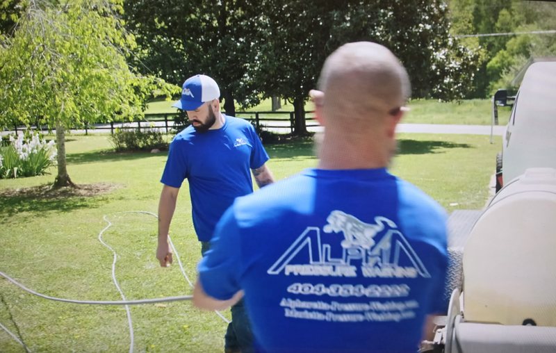 Two Alpha Pressure Washing crew members in blue company shirts working beside the equipment trailer and hoses on a lawn