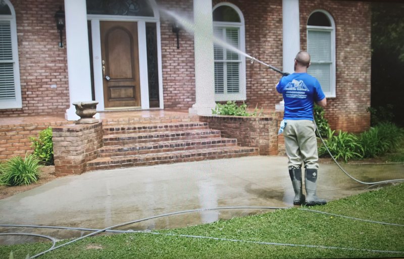 Alpha Pressure Washing technician in a blue company shirt and waders pressure washing the brick steps and concrete walkway at the front entry of a brick house